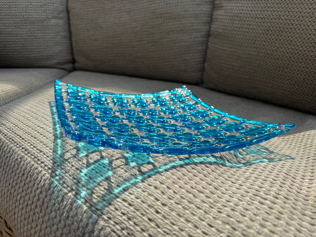 A translucent blue glass bowl with a textured geometric pattern, resting on a gray textured fabric sofa.