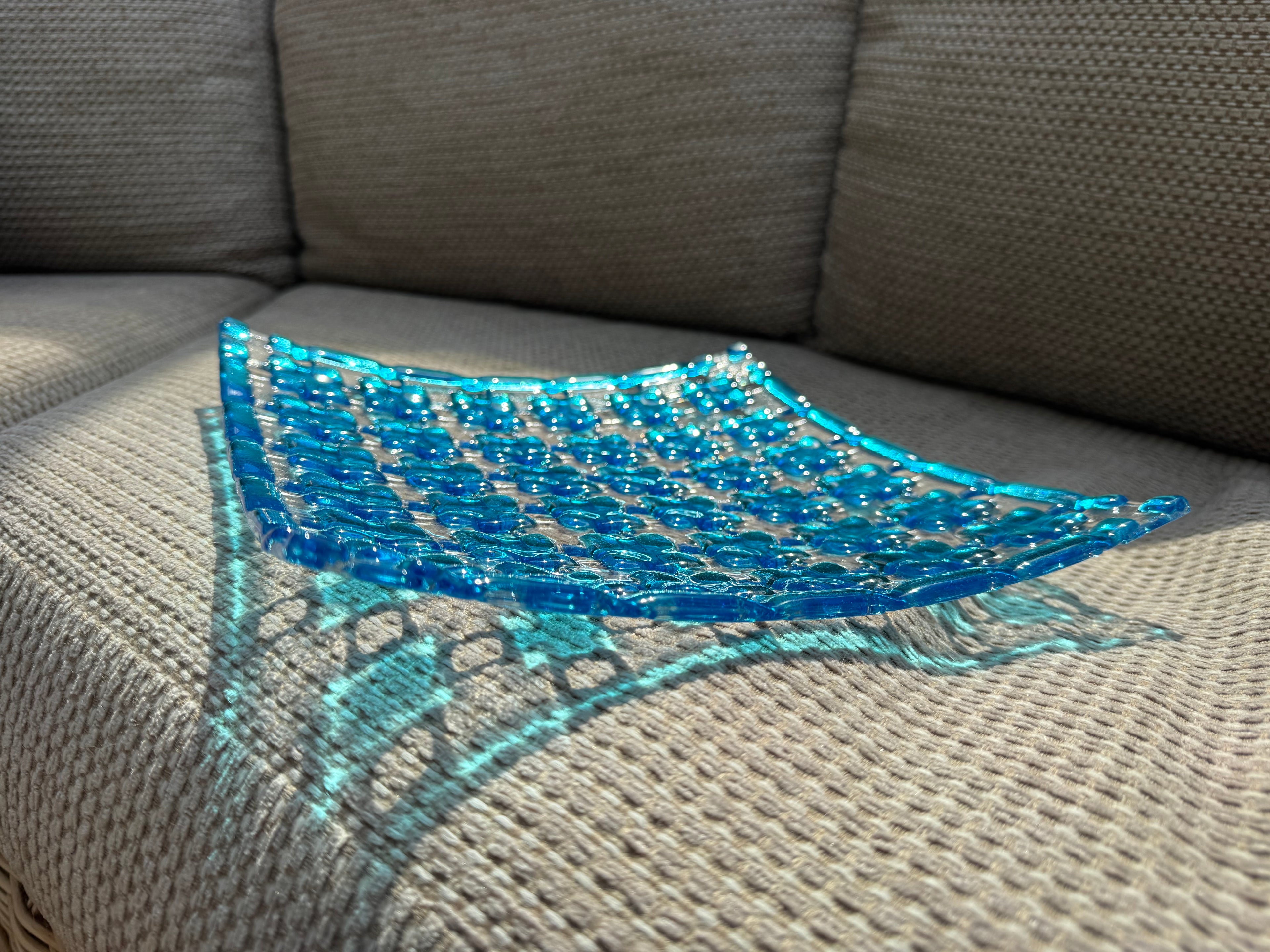A translucent blue glass bowl with a textured geometric pattern, resting on a gray textured fabric sofa.